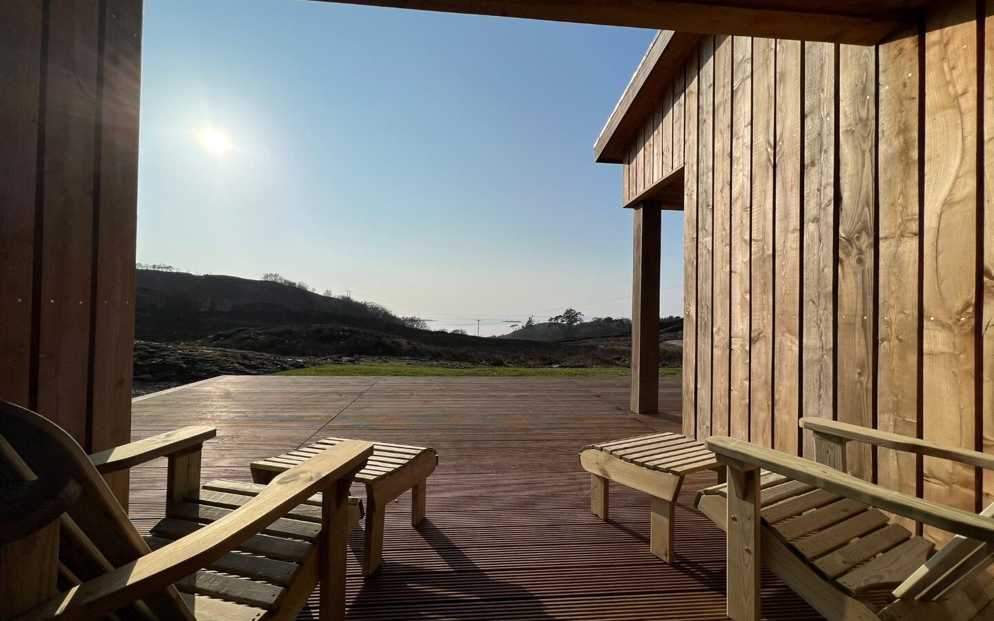 A wooden deck with two lounge chairs is bathed in sunlight, facing an open view of a clear blue sky and distant hills. The scene at Cairn Lodges is framed by the wooden exterior of a building, creating a serene and peaceful atmosphere.