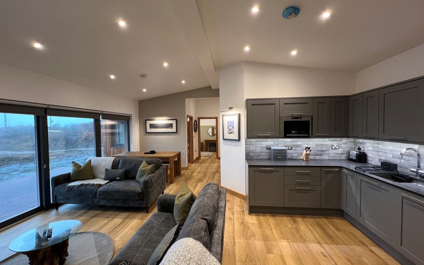 A modern open-plan living space with large windows, wooden floors, and recessed lighting. The room features a grey L-shaped sofa, a glass coffee table, and invites the feel of luxury lodges with its view into a kitchen area showcasing grey cabinets, stainless steel appliances, and a dining area in the background.