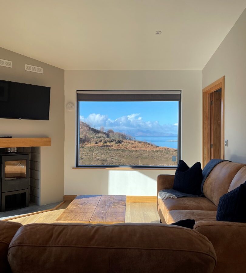 A cozy living room in one of the charming lodges, with a large window offering a scenic view of a mountainous landscape under a partly cloudy sky. The room features a brown leather sofa, a wooden coffee table, a wall-mounted TV, and a fireplace under a mantle. Light streams in through the window.