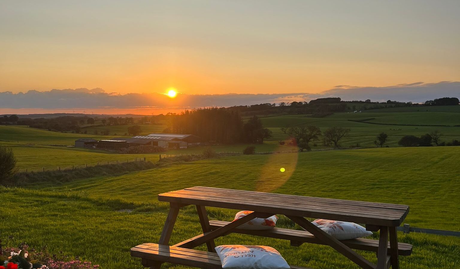 A wooden picnic table with two cushions is set on a lush green lawn, overlooking a scenic, expansive countryside. Beyond the table, the sun is setting, casting a warm glow over the rolling hills and distant trees. Vibrant flowers bloom in the garden of this charming farm retreat in the foreground.