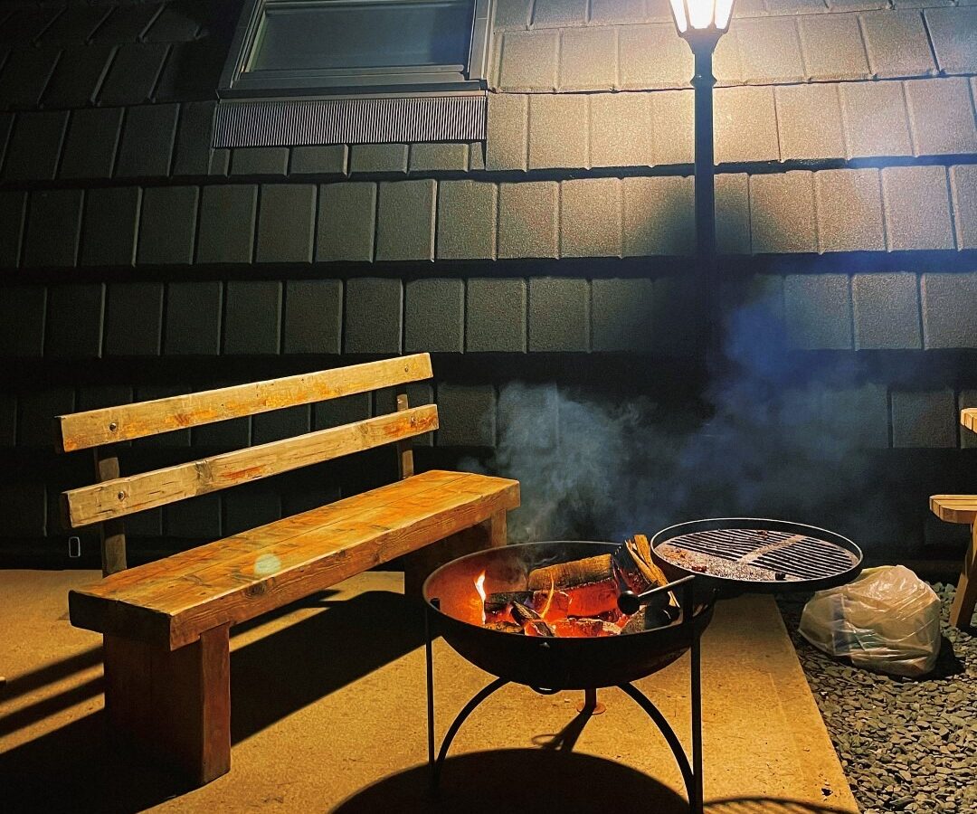 A nighttime scene features a wooden bench and a fire pit glowing with orange embers, emitting smoke. A lamp post illuminates the area, casting shadows and highlighting the shingled wall of a farm retreat in the background with a window.