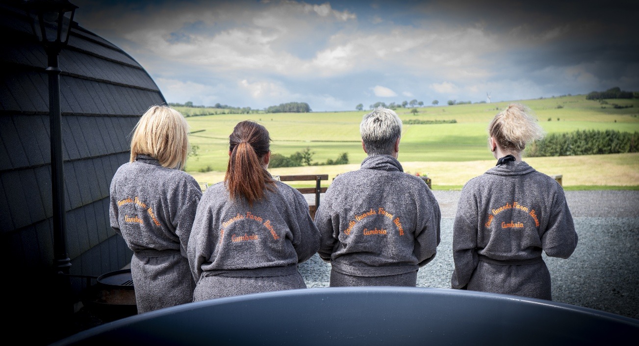 Four people wearing grey bathrobes with orange embroidery on the back stand with their backs facing the camera. They overlook a scenic landscape of green fields and, in the distance, a majestic castle retreat under a partly cloudy sky.