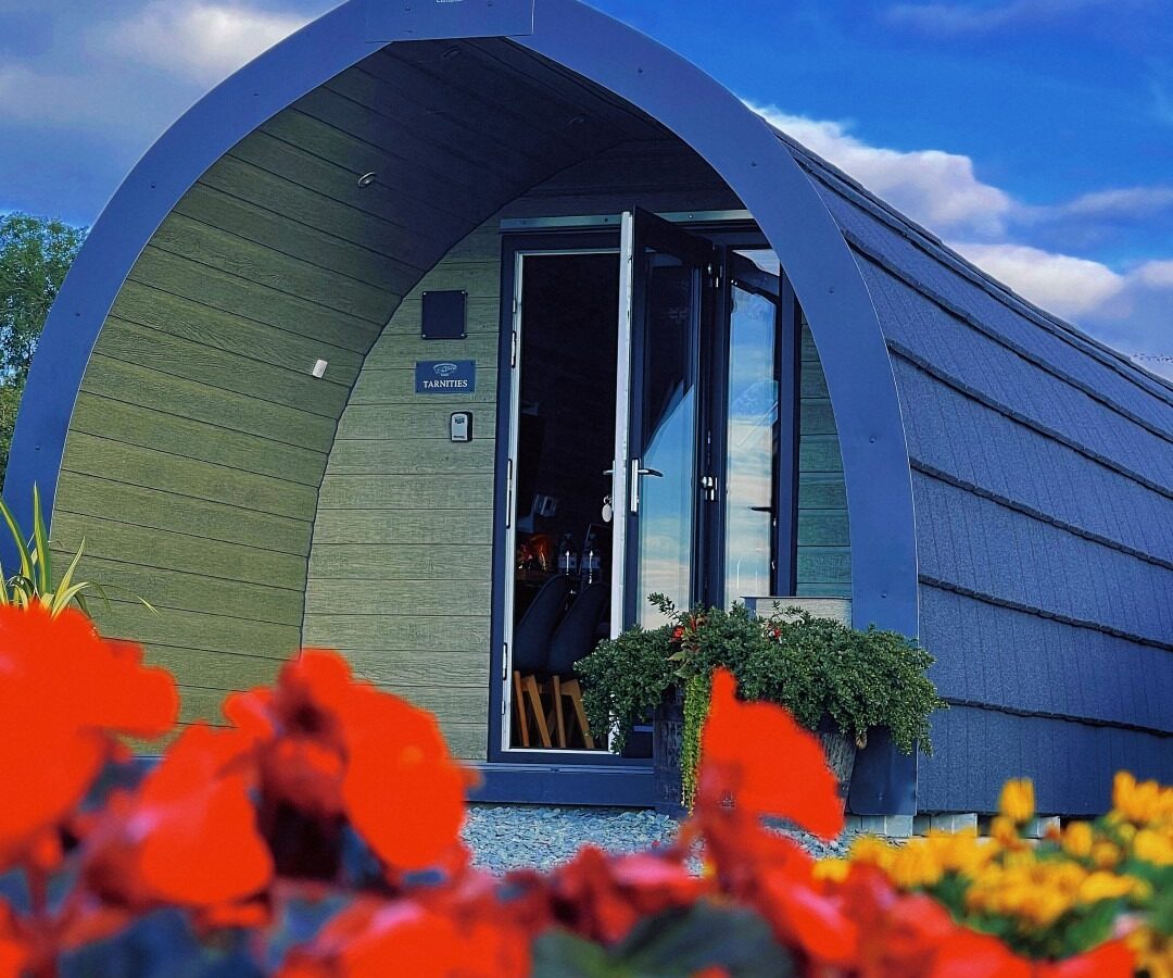 A small, modern, curved-roof cabin with wooden panels and a glass door, surrounded by vibrant red, yellow, and green flowers in the foreground. The sky is a bright blue with scattered clouds, making it an idyllic farm retreat.