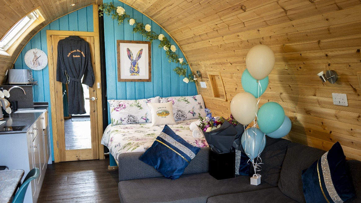 A cozy wooden cabin interior with blue accents features a bed adorned with floral bedding and plush pillows, a gray sofa with blue pillows, and a kitchen area. Balloons and a bouquet of flowers add a celebratory touch to this farm retreat. A bathrobe hangs on a door at the back.