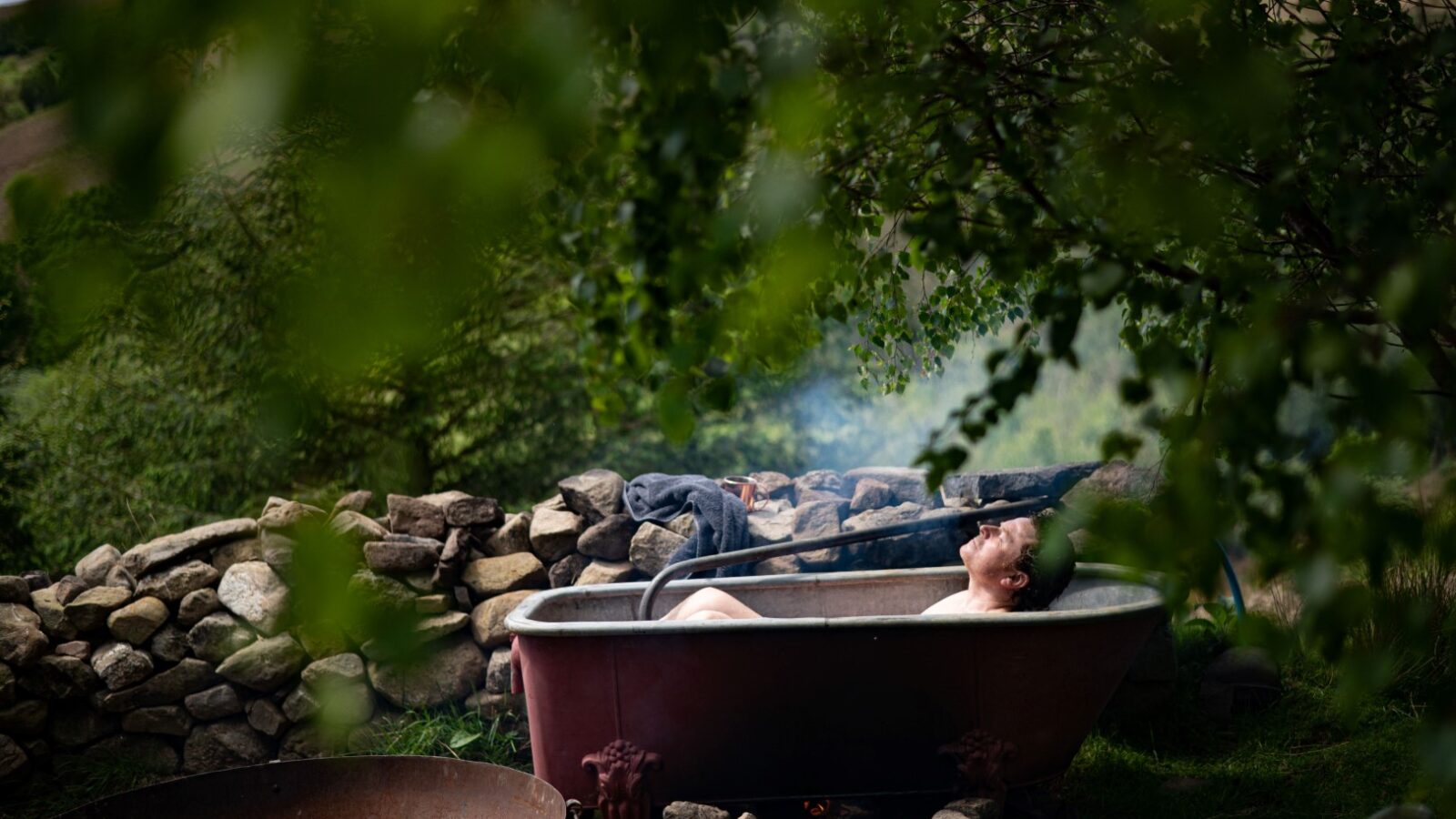 A person relaxes in an outdoor bathtub surrounded by lush greenery and a stone wall. A fire pit is in the foreground, and smoke rises, partially obscuring the person. The tranquil setting suggests a peaceful, rustic glamping experience at Red Kite’s Chillderness retreat.