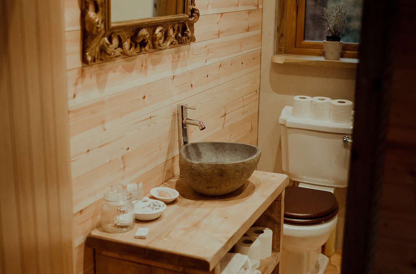 A cozy bathroom in the Netherby Treehouse boasts wooden walls and a stone sink on a rustic wooden vanity. Above the sink is an ornate gold-framed mirror. The toilet is adjacent, with a small window above it letting in natural light. Towels and toiletries are neatly arranged on the vanity shelves.