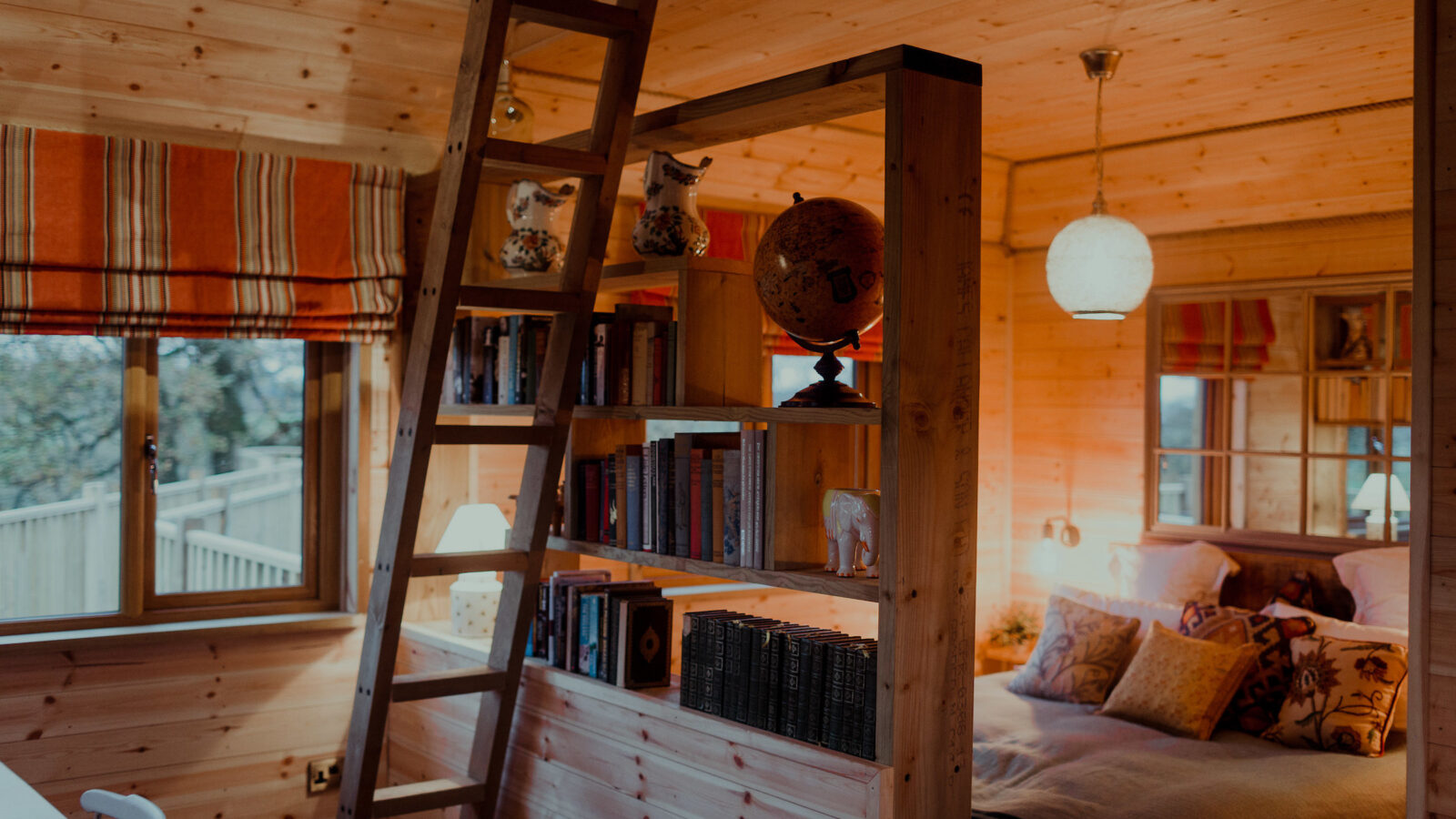 The Netherby Treehouse offers a cozy wooden cabin interior with a loft bed accessed by a wooden ladder, a bookshelf filled with various books, and unique decorative items. Warmly lit with soft lighting, the room features striped curtains and patterned pillows, making it an inviting and unique accommodation.