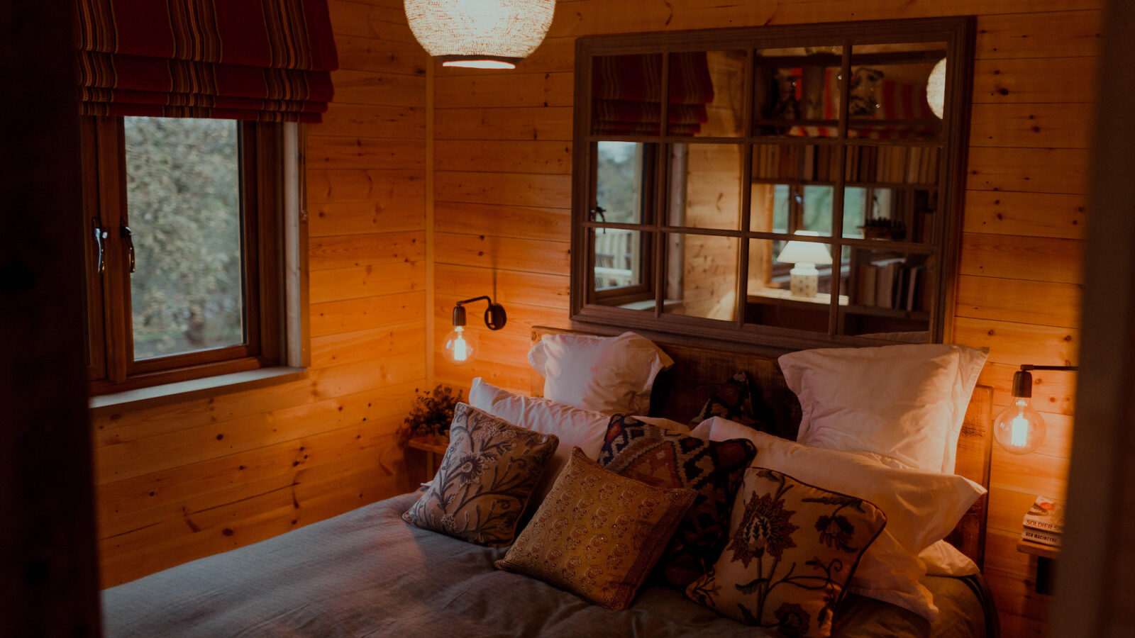 A cozy bedroom in the Netherby Treehouse features a wooden interior with a large bed adorned with decorative pillows and soft lighting. A mirror above the bed reflects the room, adding depth. Windows with striped curtains let in natural light, enhancing the warm, inviting atmosphere.