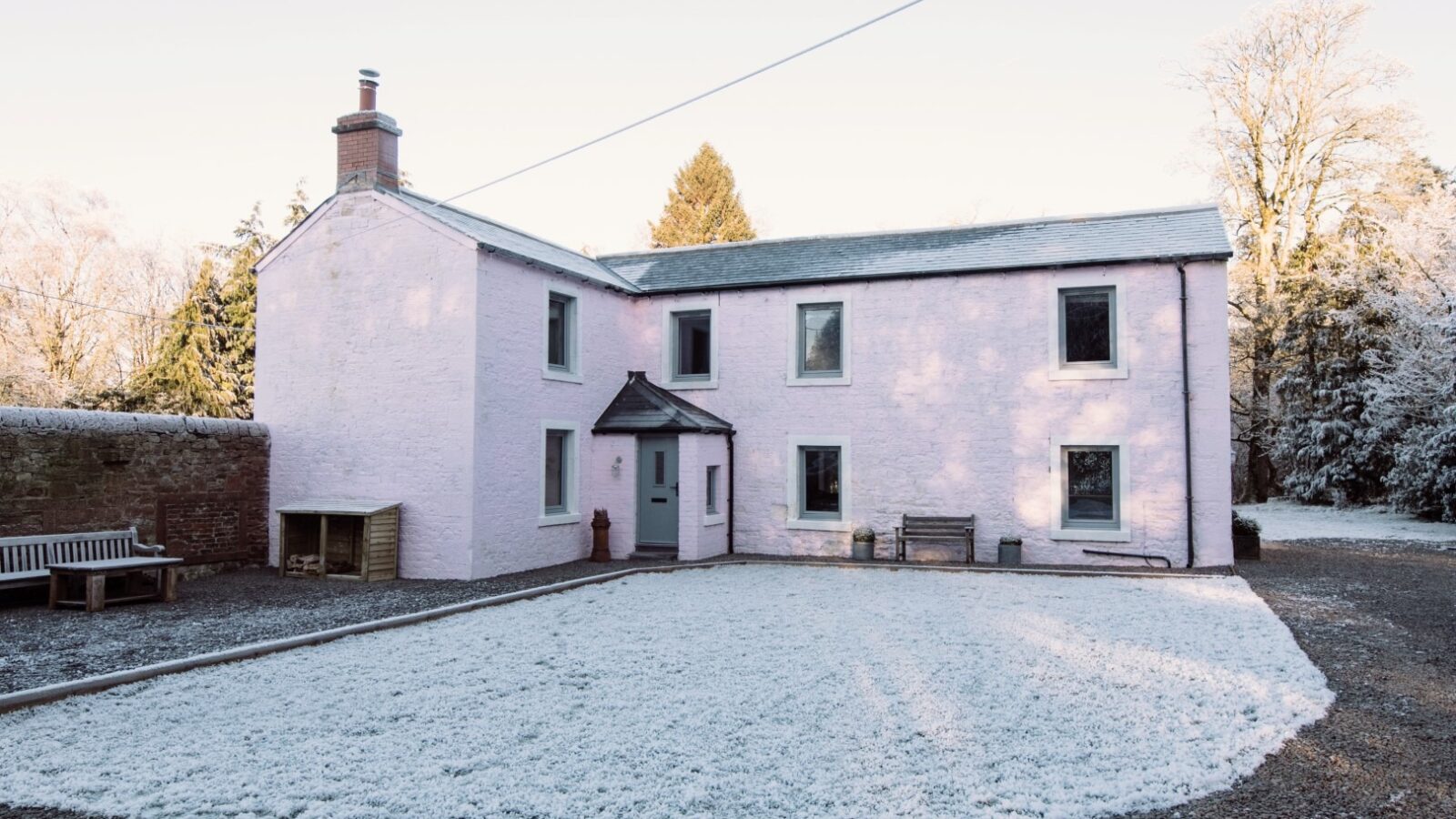 A two-story pink house with several windows stands in a snowy yard at Cleugh Foot. The house has a chimney and a small wooden shelter next to a door on the left. A bench is placed against the front wall on both sides, and trees can be seen in the background.