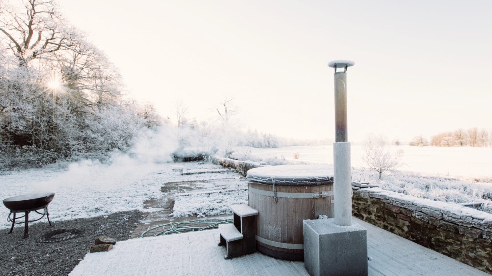 A snowy outdoor scene at Cleugh Foot shows a wooden hot tub with a metal chimney on a wooden deck next to a stone wall. A small set of steps leads to the hot tub. In the background, there's a metal fire pit emitting smoke. Bare trees and a snow-covered field are visible.