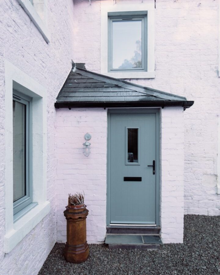 A quaint light-pink brick house at Cleugh Foot features a grey front door with a window. There is a modern wall light beside the door and a small stone step leading to it. An old wooden barrel planter filled with sticks sits next to the entrance on a gravel surface.