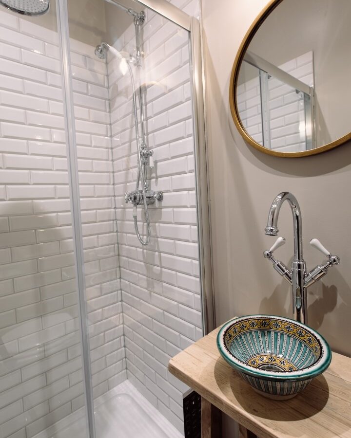 A modern bathroom at Cleugh Foot features a glass-enclosed shower with white subway tiles. A wooden vanity holds a decorative, multicolored ceramic sink with a silver faucet. Above the sink is a round mirror with a gold frame on a beige wall.