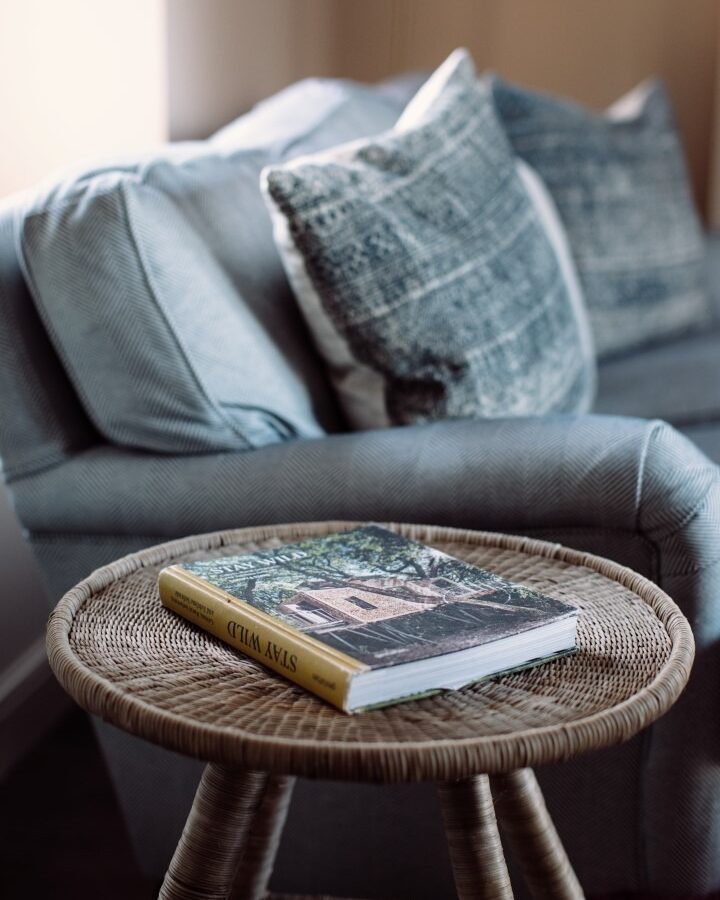 A cozy seating area at Cleugh Foot features a light blue, textured sofa adorned with matching cushions. In the foreground, a round wicker side table holds an open book with a yellow spine and a picturesque cover. Soft light filters in, creating a warm and inviting atmosphere.