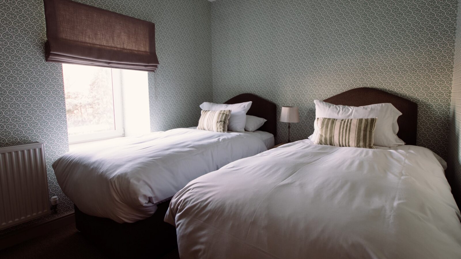 A cozy bedroom at Cleugh Foot with two single beds, each with white bedding and a pillow with a striped cover. The room has a window with a brown blind, an off-white radiator beneath the window, and patterned wallpaper in soft colors. A small lamp is placed between the beds.