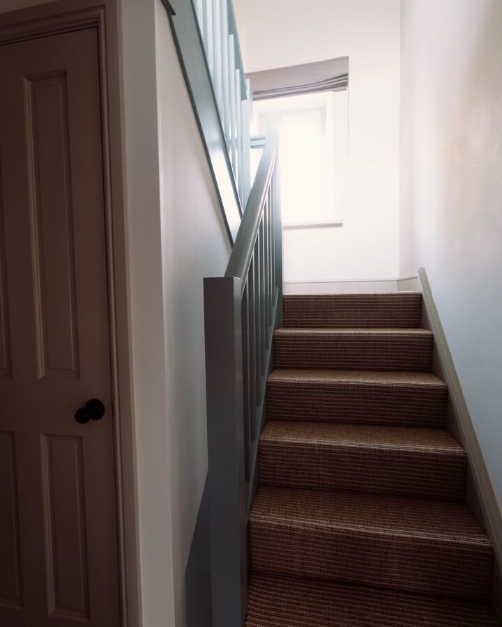 A staircase with beige carpeted steps leads up to a brightly lit landing with a window at the top. The Cleugh Foot staircase is bordered by a blue-gray railing, and a closed wooden door with a black knob is on the left side at the bottom of the stairs.