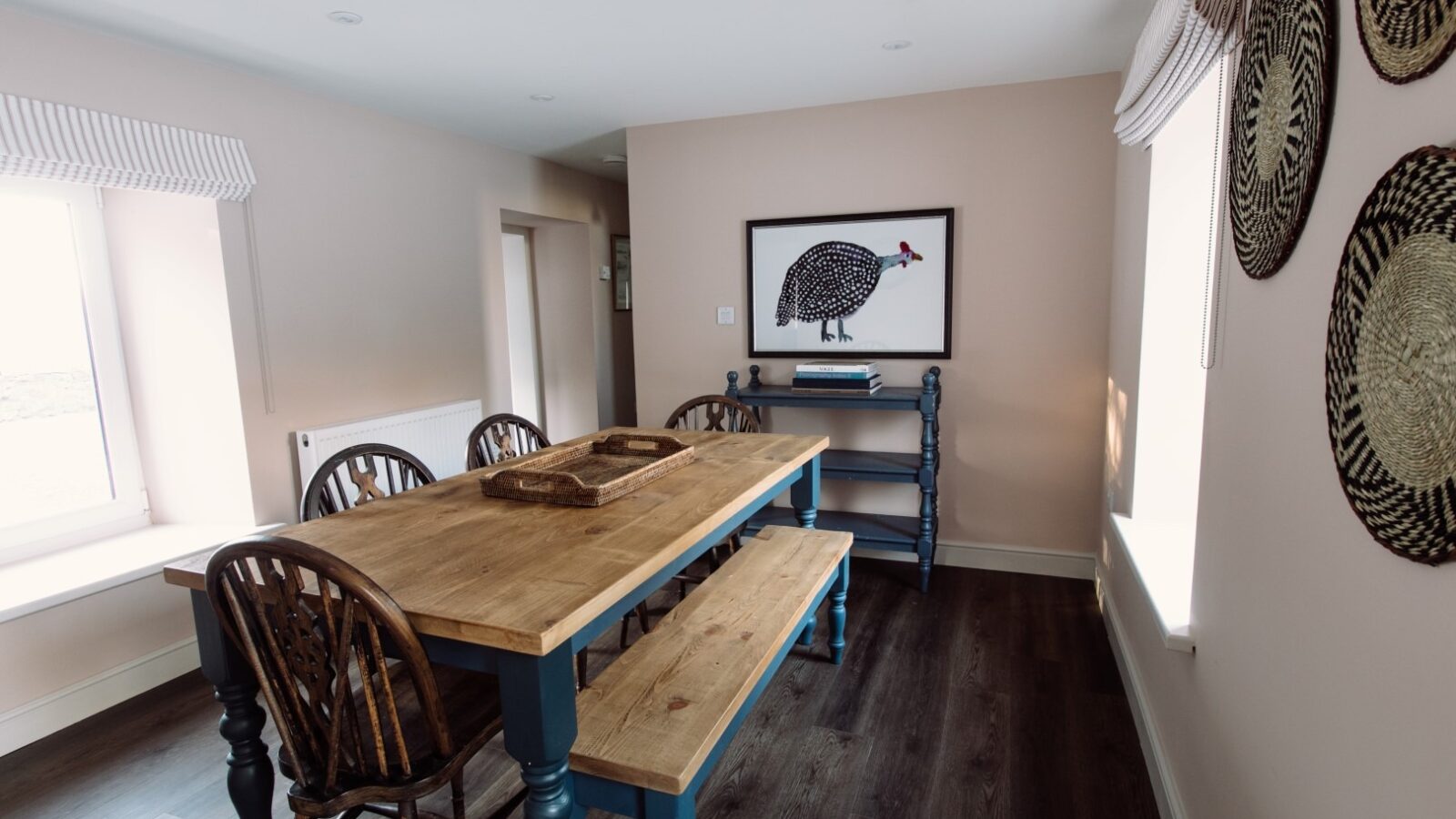 A bright dining room with a wooden table and benches, complemented by matching chairs. The light walls are adorned with woven basket wall hangings and a framed bird print. Large windows on two walls let in natural light, revealing Cleugh Foot-inspired dark hardwood floors.