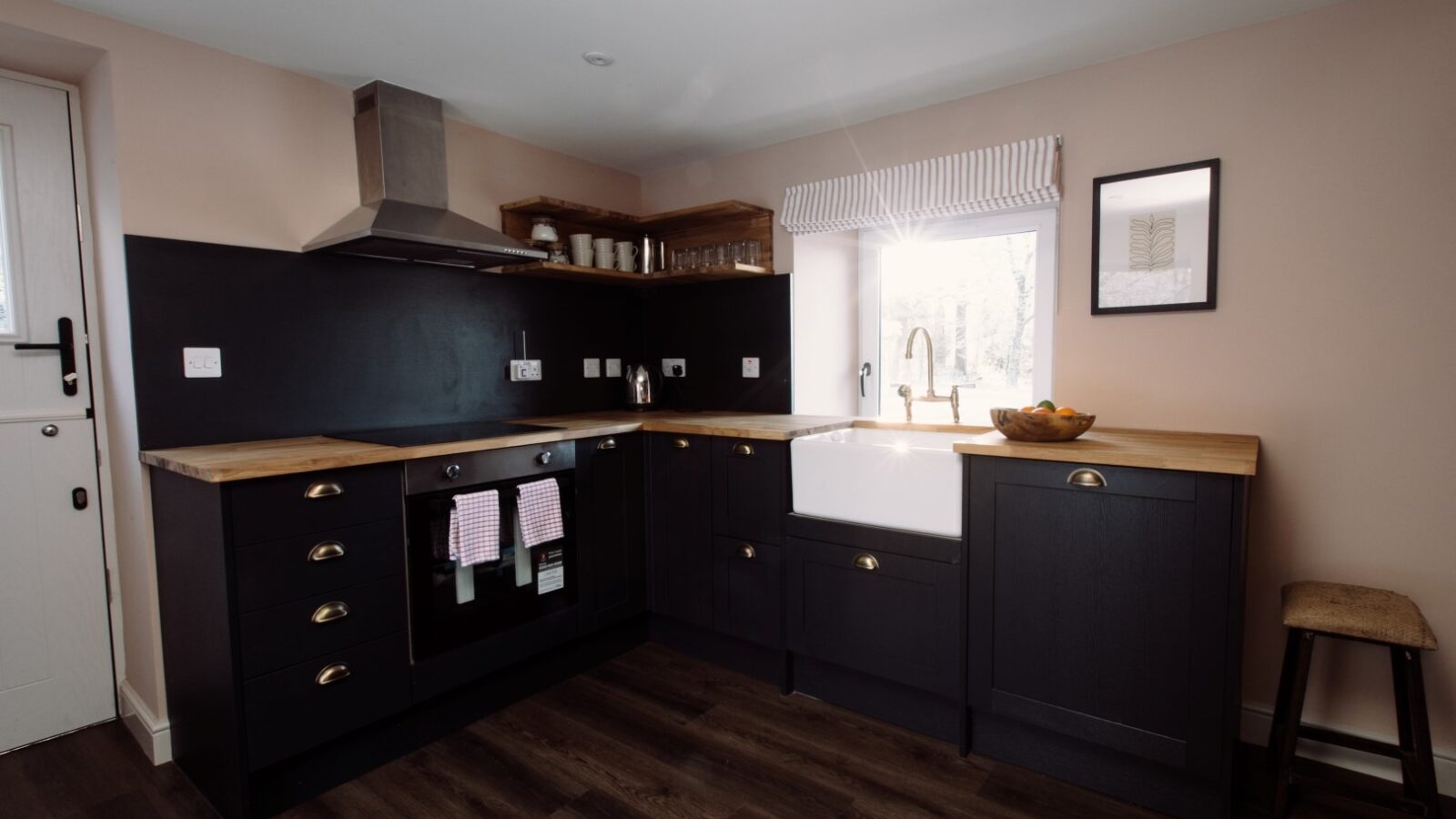 A modern kitchen with dark cabinetry, wooden countertops, and a farmhouse-style sink. There's a stainless steel stove with an overhead range hood on the left, open shelves with dishes above, a window with a striped valance from Cleugh Foot, and a stool under the countertop.