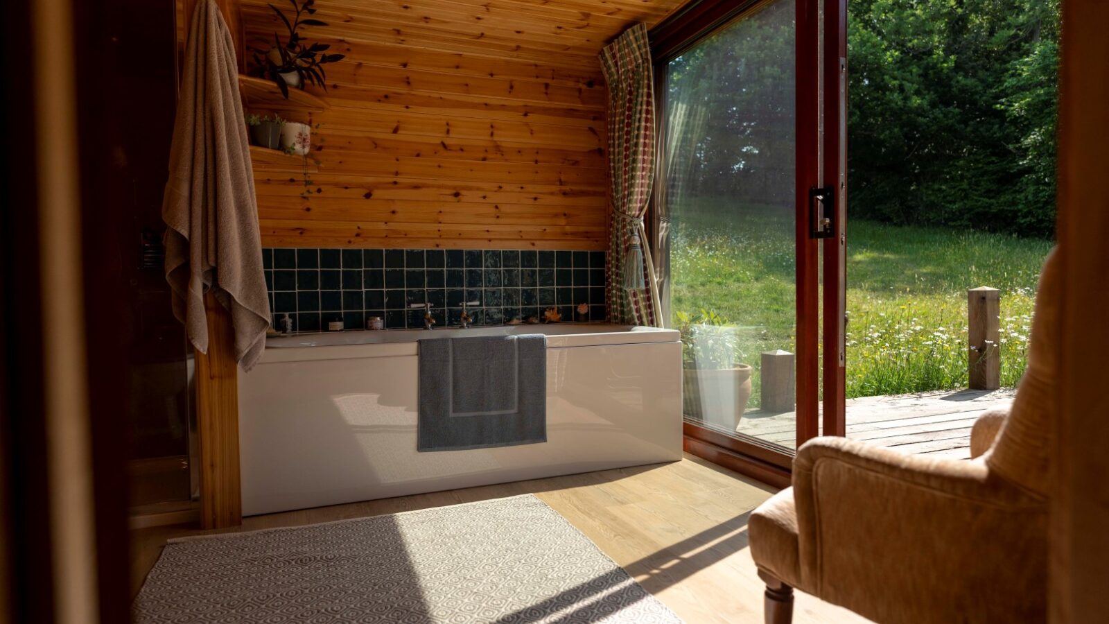 A cozy bathroom at Cobnut Cabin features wooden walls and ceiling, showcasing a bathtub with a green tile backsplash. A towel hangs over the edge of the tub. Sunlight streams in through large sliding glass doors that open to a lush, grassy outdoor area. An armchair sits nearby, perfect for unwinding at this Ritty Retreats getaway.