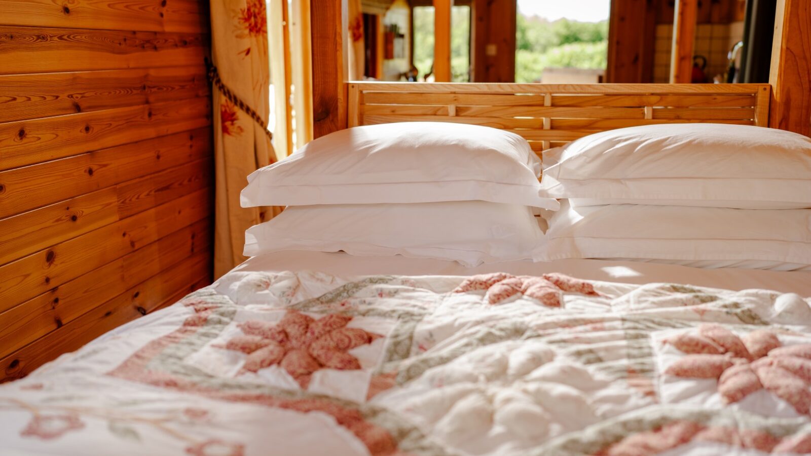 A cozy bed with a floral quilt and four neatly stacked white pillows, set in Cobnut Cabin. Sunlight streams in through a window, creating a warm and inviting atmosphere, perfect for your serene cabin retreat with Ritty Retreats.