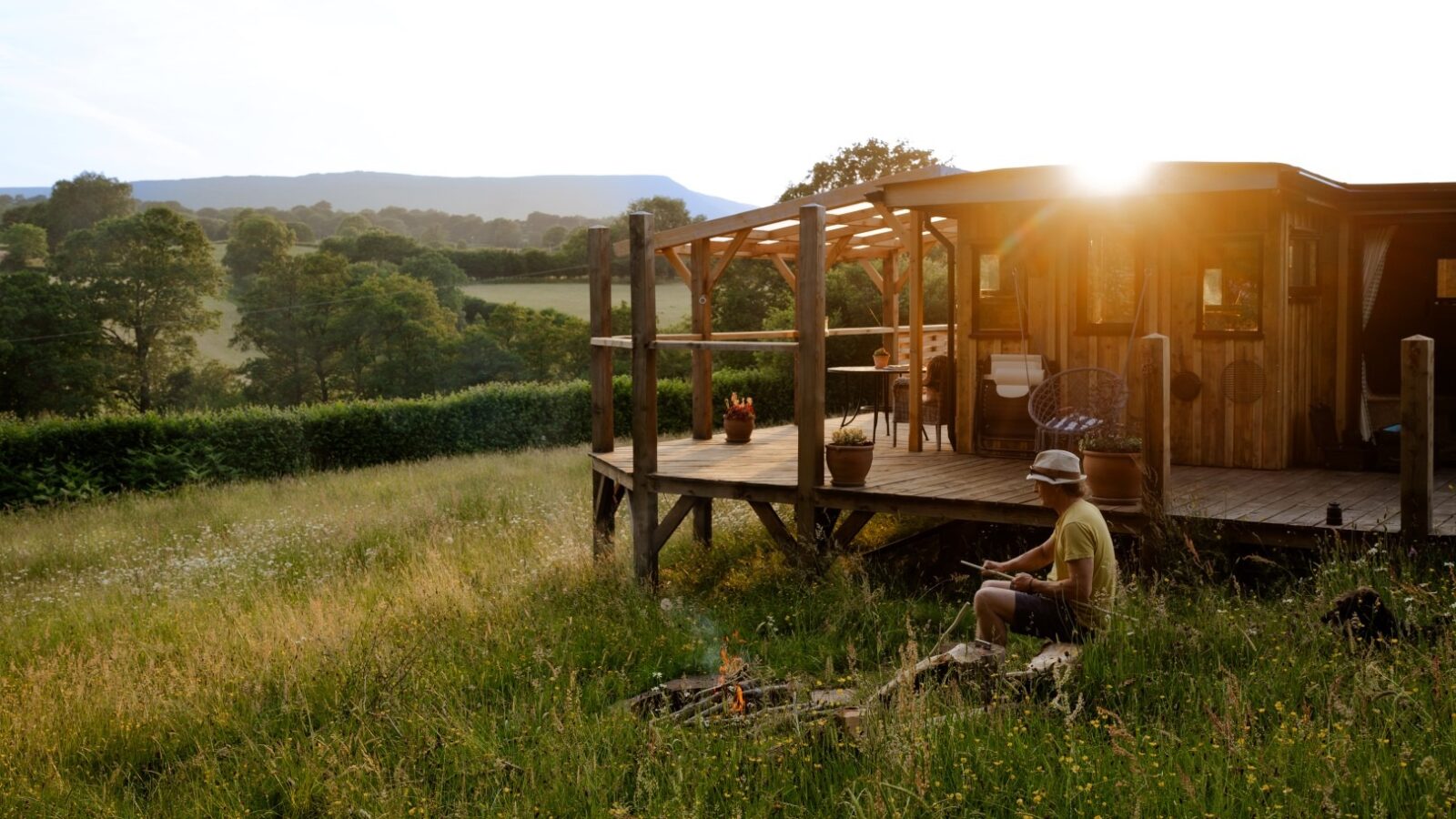 A person is sitting in front of a fire pit in a grassy field near Cobnut Cabin, an offering from Ritty Retreats, with a large deck. The sun is setting behind the distant hills, casting warm light over the landscape. Trees and bushes surround the cabin rentals, contributing to the peaceful atmosphere.