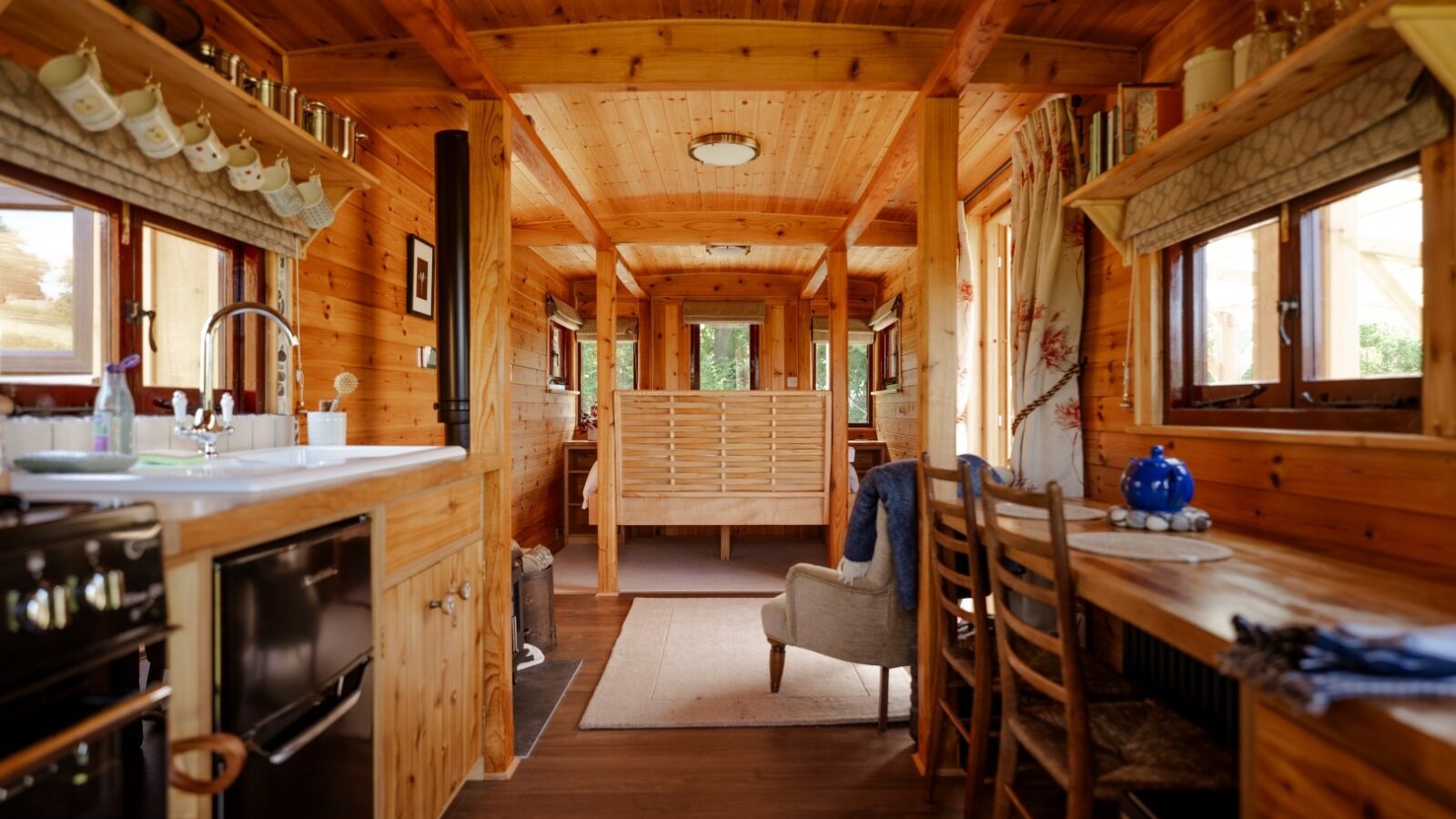 The interior of the cozy Cobnut Cabin features a rustic design. On the left, there's a compact kitchen with a stove and sink. A wooden table with chairs sits on the right, leading to a sitting area and bed in the background. Multiple windows allow natural light to stream into this charming cabin retreat.