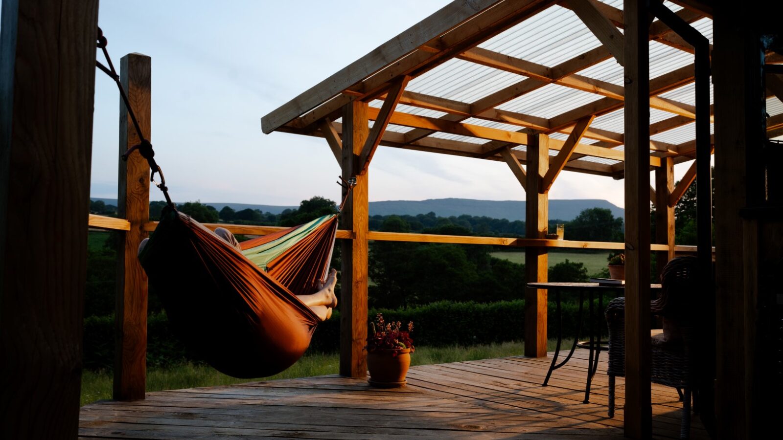 A wooden deck at Cobnut Cabin, part of the Ritty Retreats collection, featuring a pergola with a hanging hammock on the left and a potted plant. Beyond the deck, enjoy a scenic view of green hills and trees under a sky transitioning to dusk. A wicker chair and table grace the right side.