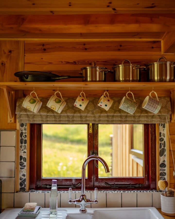 A cozy wooden kitchen in Cobnut Cabin features a double sink under a window. Above the sink, cups with colorful patterns hang from hooks beneath a shelf holding pots and pans. On the sink counter, there's a bottle, a sponge, and a soap dish. Natural light illuminates this peaceful cabin retreat scene.