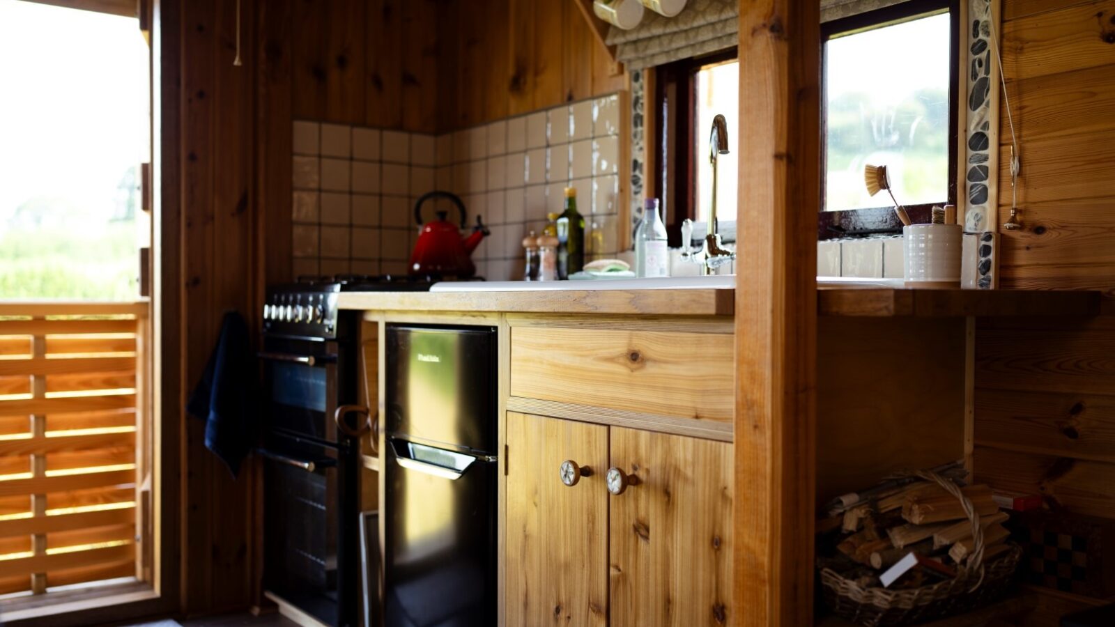 Cozy wooden kitchen at Cobnut Cabin with a small refrigerator, gas stove, sink, and tiled backsplash. Kitchen utensils and condiments are placed on the counters. A window above the sink lets in natural light, and a door with a wooden slat design is partially open on the left.