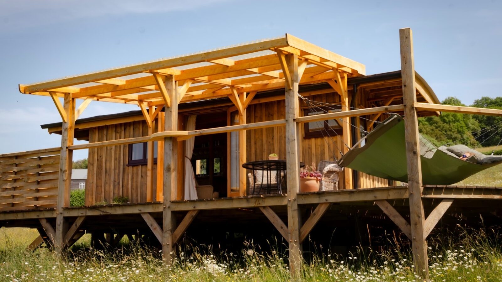 A small wooden cabin, dubbed the Cobnut Cabin by Ritty Retreats, features a large deck with a pergola. On the deck, you'll find a table with chairs and a hammock strung up on the right side. The charming cabin retreat is surrounded by a grassy field adorned with wildflowers and trees in the background.