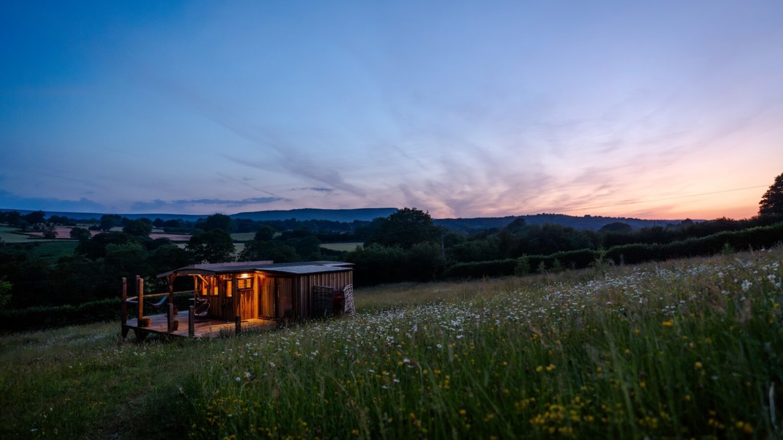 A small, lit Cobnut Cabin sits in the middle of a field of grass and wildflowers at dusk. The sky is transitioning from blue to shades of pink and purple as the sun sets behind the distant hills. Trees and hedges surround the field, enhancing the serene countryside atmosphere typical of Ritty Retreats.