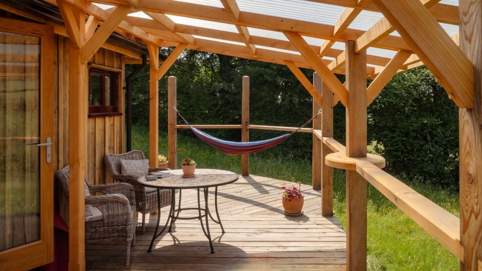 A wooden patio with a transparent roof features two wicker chairs and a round table with a small potted plant. A colorful hammock is hung in the background, and another potted plant sits on the deck. The patio at Cobnut Cabin overlooks a grassy yard and greenery, providing an ideal spot for cabin retreats.