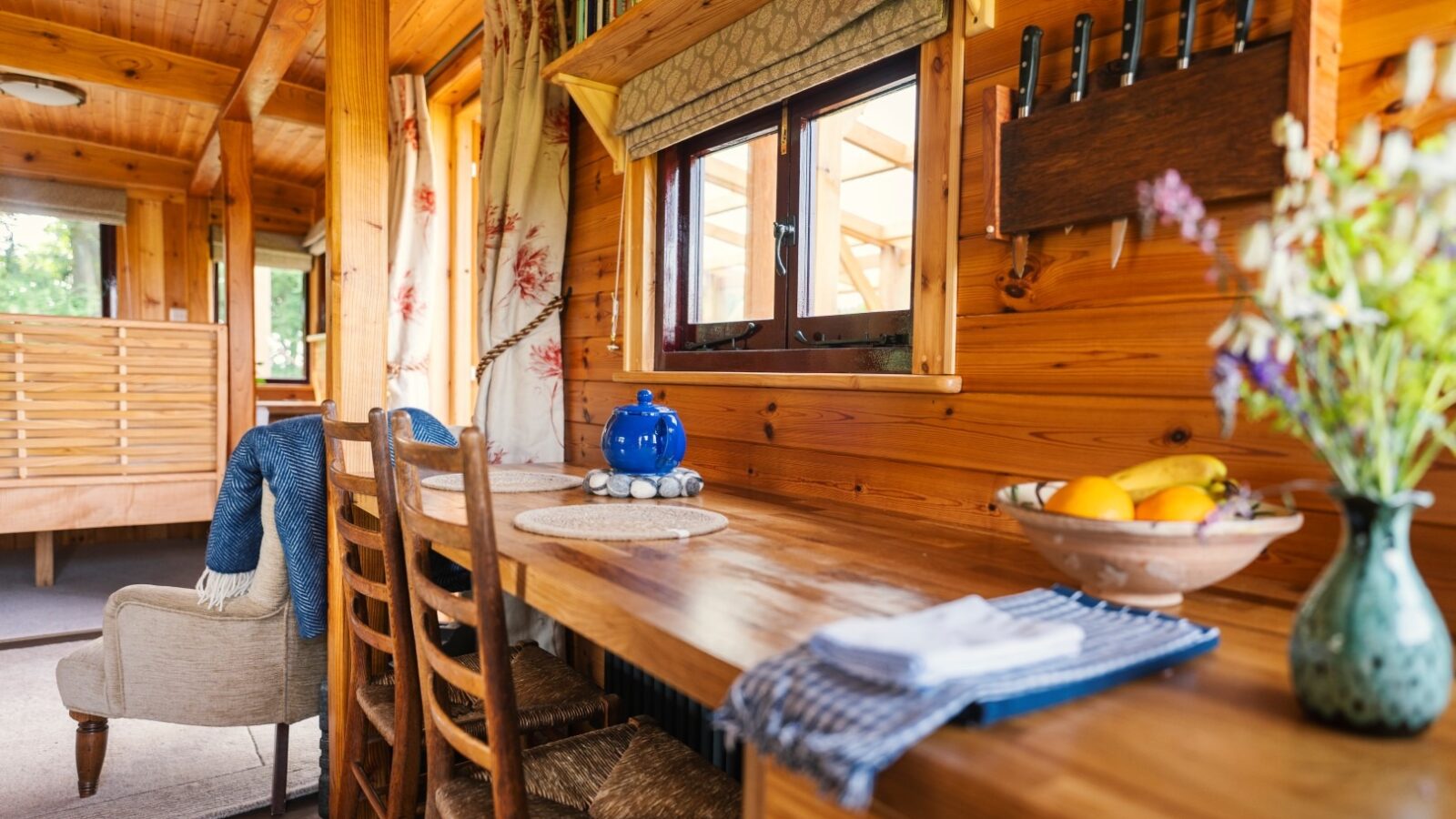 Cozy wooden interior of a small home with a long wooden table and chairs near the window. The table is adorned with a blue teapot and a bowl of fruits. A vase with flowers and cutlery are in the foreground, while a cushioned chair draped with a blanket adds to the Ritty Retreats charm.