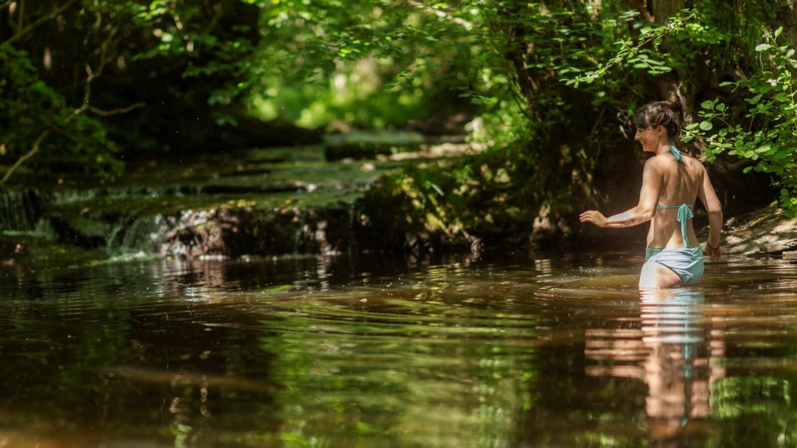 A person in a bikini wades through a calm forest stream, surrounded by lush greenery. Sunlight filters through the trees, illuminating the serene scene. A small waterfall is visible in the background, not far from the cozy haven of Ritty Retreats.