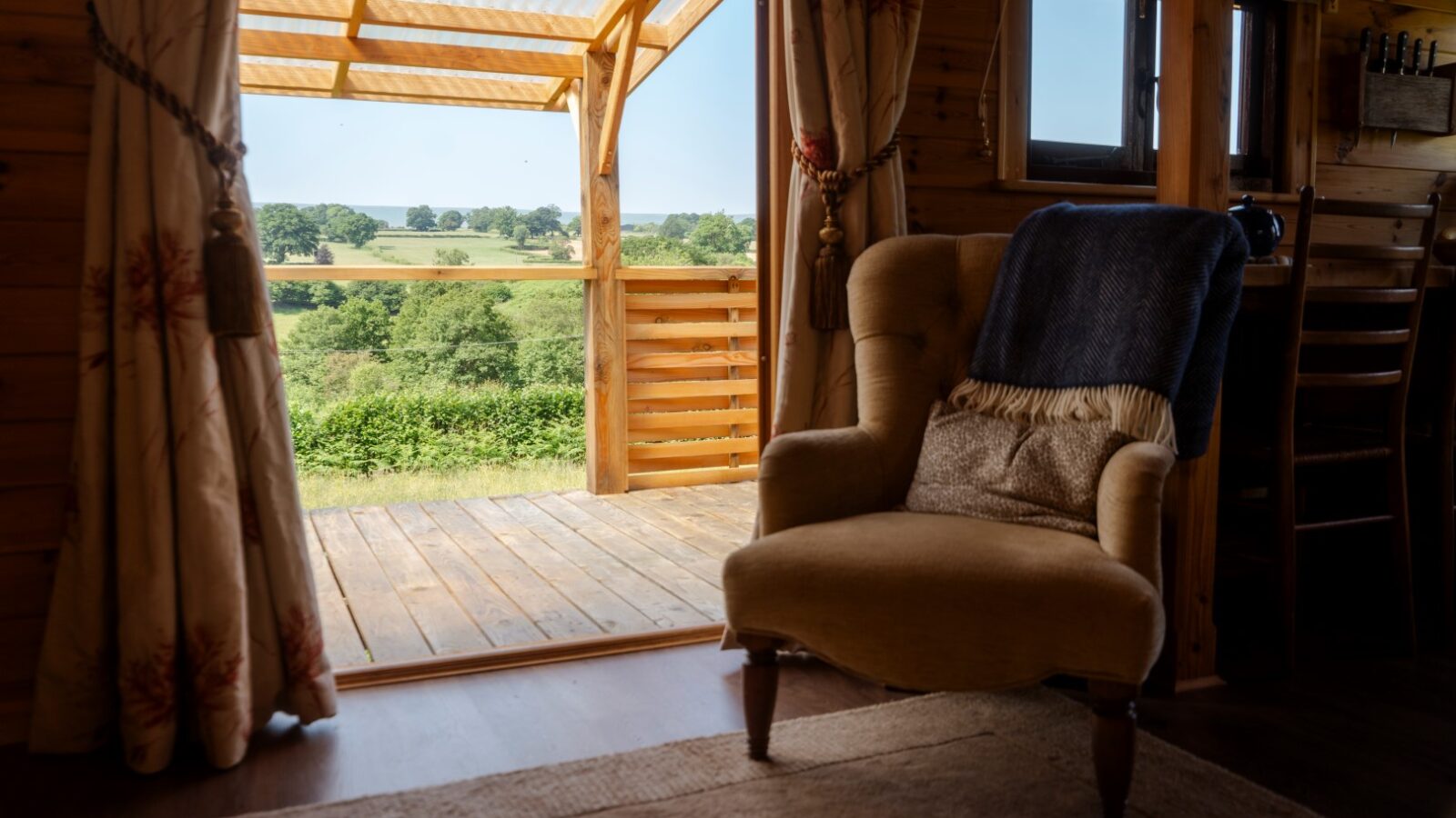 A cozy wooden cabin interior at Cobnut Cabin features an armchair draped with a blanket beside an open door leading to a small deck. The deck overlooks lush, green countryside with fields and trees under a clear blue sky. Cream curtains frame the door, making it one of the finest cabin rentals by Ritty Retreats.