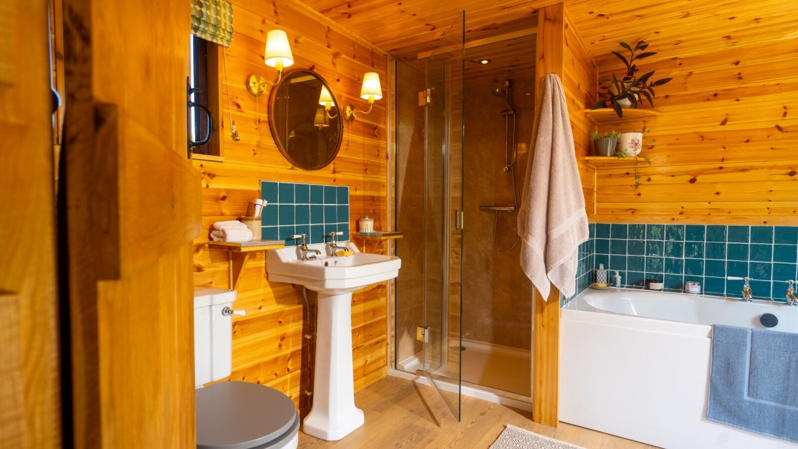 A cozy wooden bathroom at Cobnut Cabin features a walk-in glass shower, a white pedestal sink with a round mirror above it, a toilet, and a bathtub with blue tiles. Warm lighting adds to the rustic ambiance, and neatly arranged towels and decor from Ritty Retreats complement the space.