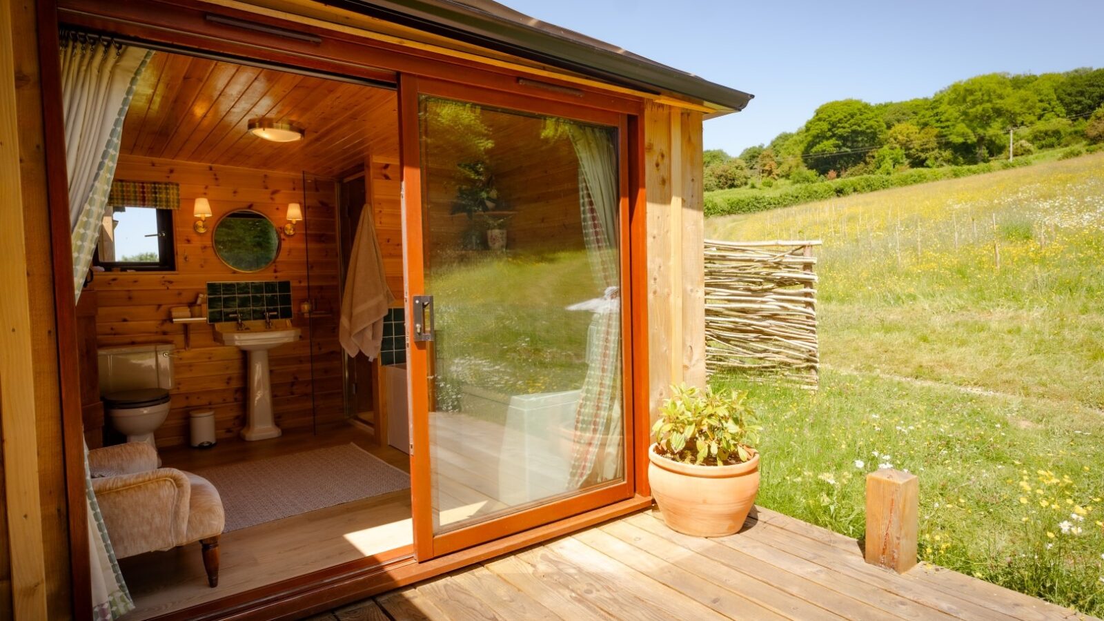 A cozy wooden bathroom in the Cobnut Cabin features a large sliding glass door that opens onto a sunny deck. Inside, there's a sink, mirror, and toilet with a towel hanging on the wall. Outside, a potted plant sits by the door as the cabin rental overlooks a lush meadow with trees in the background.