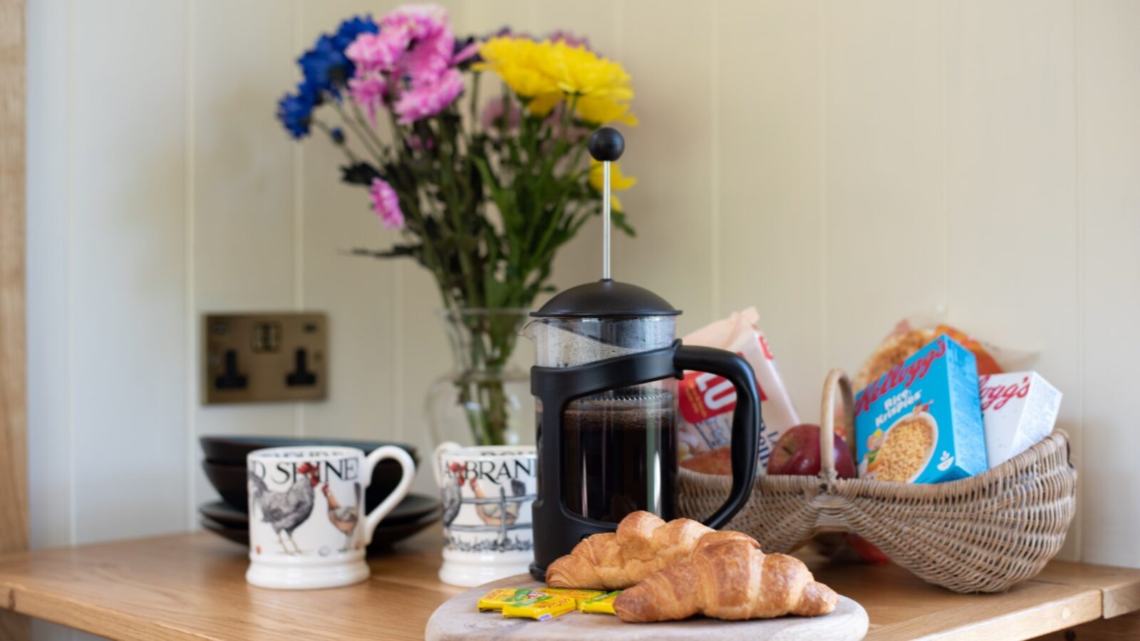 A wooden countertop with a French press filled with coffee, two croissants on a wooden board, a packet of butter, a basket with cereal boxes, two mugs with a rooster design featuring shepherd huts in the background, and a vase with colorful flowers.