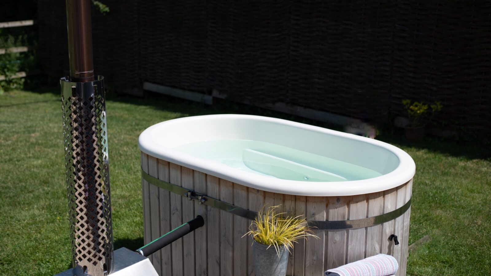 A wooden hot tub filled with water is set on green grass in a cozy backyard, reminiscent of tranquil shepherd huts. Rolled white towels with colorful stripes sit on a small stool next to the hot tub, and a plant in a pot adds charm. A tall metal chimney is attached for heating.