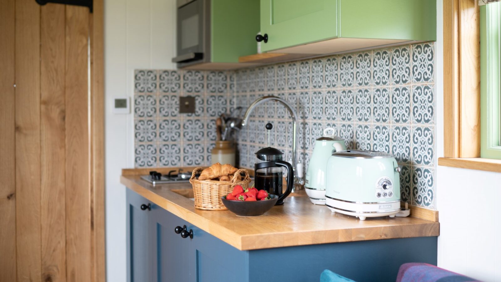 A cozy kitchen with green and blue cabinets, wooden countertops, and patterned backsplash tiles. A basket with croissants, a bowl of strawberries, a French press, a mint green kettle, and a toaster are placed on the counter. A wooden door is partially visible on the left. The warmth feels like stepping into one of those charming shepherd huts.