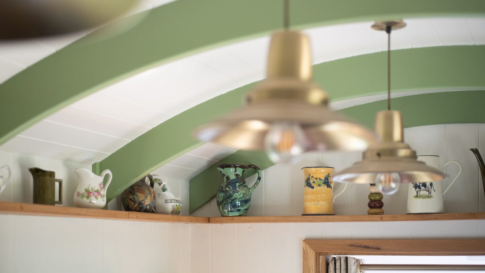 A shelf mounted near the ceiling displays a collection of vintage pitchers and jugs in various colors and patterns. The scene, reminiscent of cozy huts, is illuminated by pendant lights with large, metallic shades. The ceiling features green arch beams contrasting with white paneling.