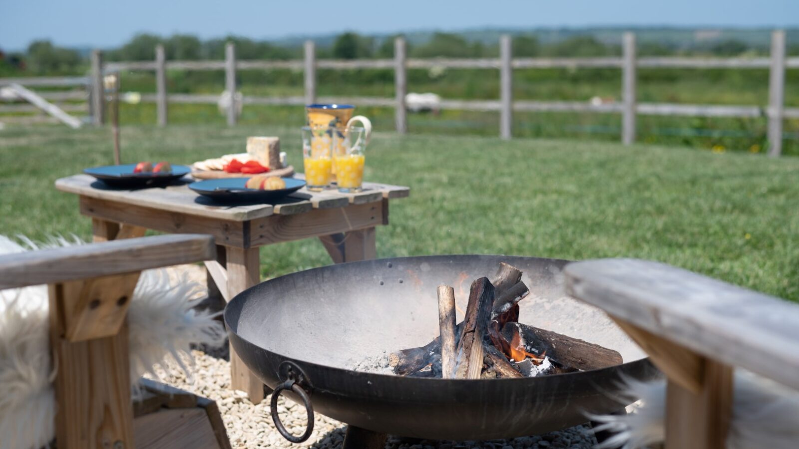 Outdoor scene featuring a fire pit with burning logs on a gravel surface, reminiscent of cozy gatherings by rustic shepherd huts. Surrounding the fire pit are wooden benches with white cushions. Picnic food, including bread, cheese, and watermelon, along with a pitcher of orange juice, is set on small wooden tables.