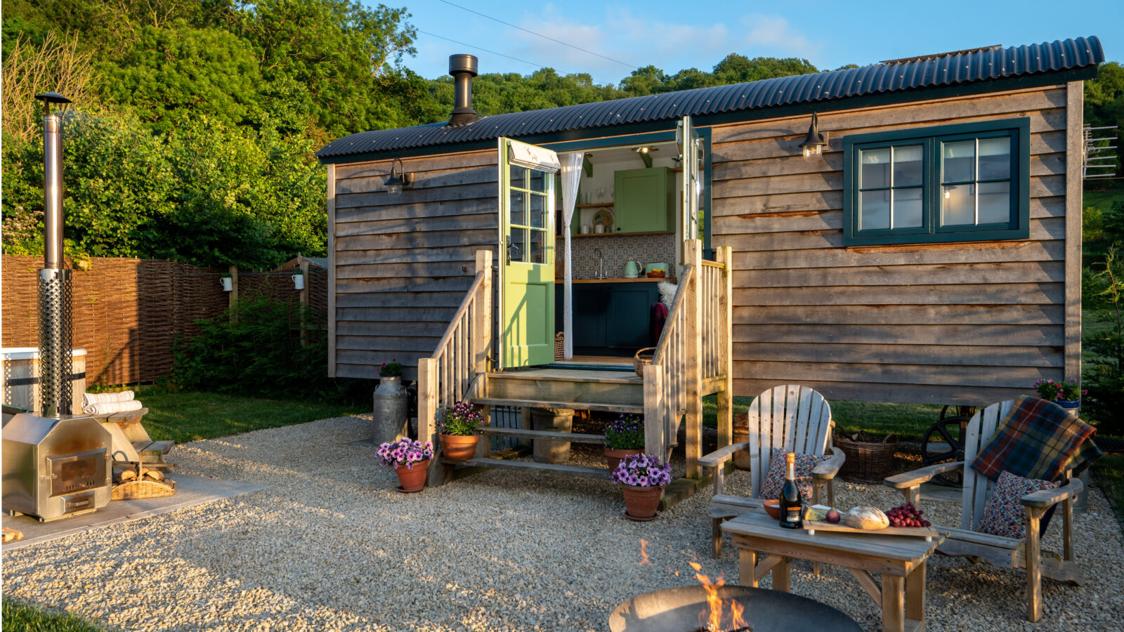 A cozy, rustic tiny house with a wooden exterior, partially open green doors, and a small outdoor area. The front yard features Adirondack chairs, potted plants, a fire pit with crackling flames, and a small outdoor oven. Green trees and scattered clouds frame the scene like picturesque Shepherd Huts in the countryside.