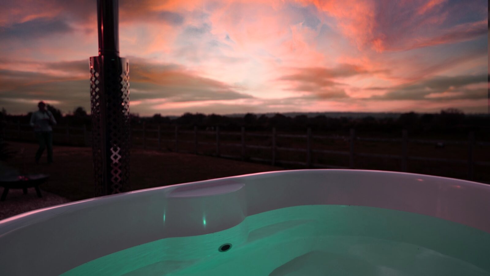 Hot tub with gently glowing water sits outdoors, with a beautiful sunset painting the sky in pink, orange, and purple hues. A metal chimney is visible to the left, near a charming Shepherd Hut, with a Collie standing in the distance near a wooden fence.