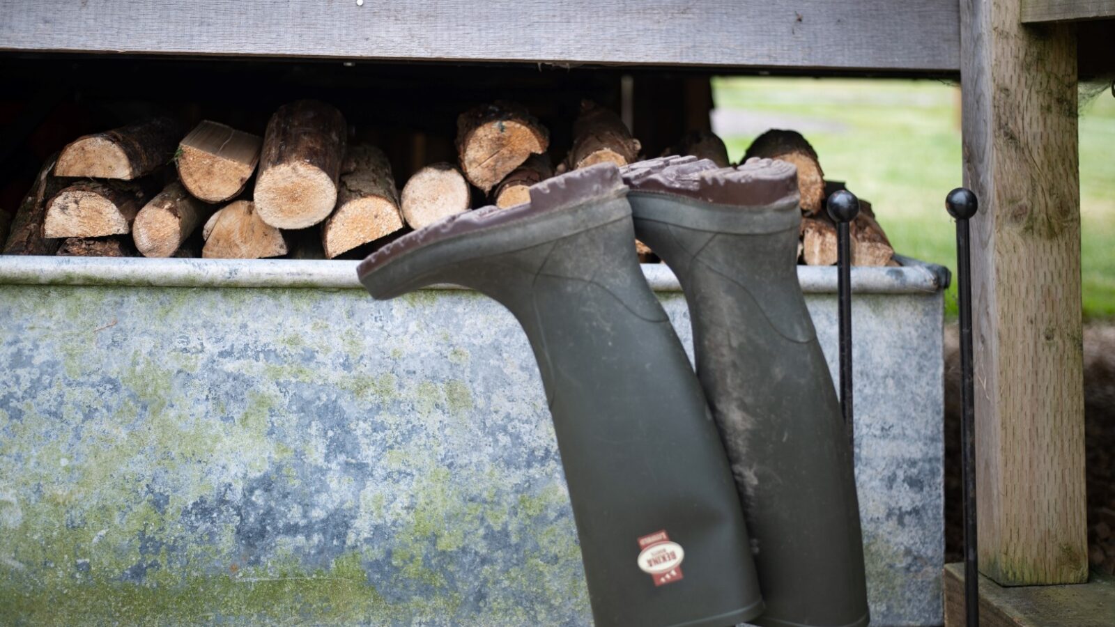 A pair of muddy green rubber boots are propped upside down on stakes in front of a metal container filled with cut firewood. Nearby, a Collie rests peacefully. The scene is set outdoors on a gravel surface, with quaint Shepherd Huts in the background.