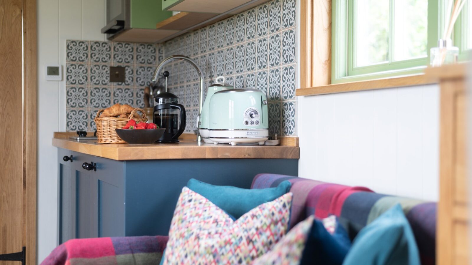 Cozy kitchen area featuring a stylish tiled backsplash, a green under-counter cabinet, wooden countertop, and mint green toaster. A basket of bread and a bowl of strawberries are on the counter. Colorful cushions and a plaid blanket adorn a nearby sofa, reminiscent of rustic shepherd huts.