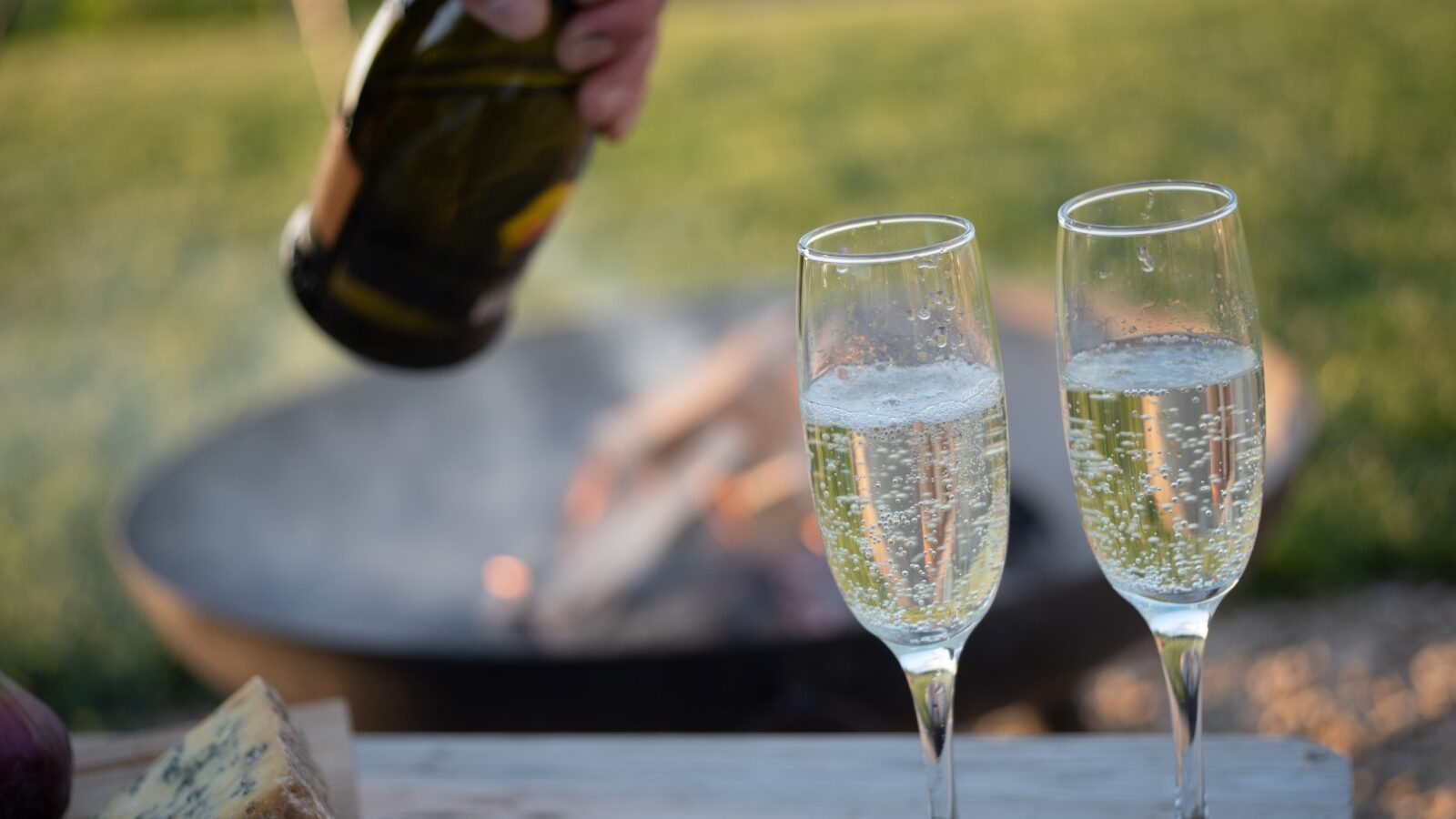 Two filled champagne flutes sit on a wooden surface outdoors, with a hand holding a champagne bottle nearby. In the background, there is a fire pit with a small fire burning, and green grass beyond. A wedge of cheese is partially visible on the table, while a Collie lounges peacefully near one of the huts.