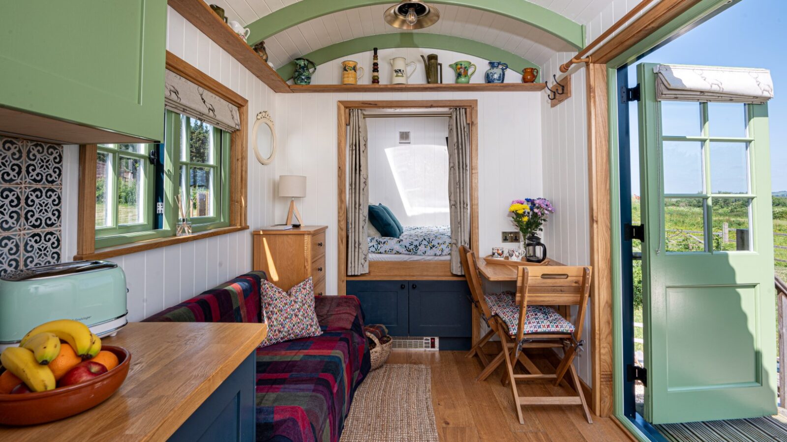 A cozy, well-lit tiny house interior with green and white accents. It has a small kitchen with a green toaster and fruit bowl, a sofa covered in a colorful blanket, a compact dining table with chairs, and a small lofted bed in the background. The open door reveals a scenic view resembling those seen from shepherd huts.