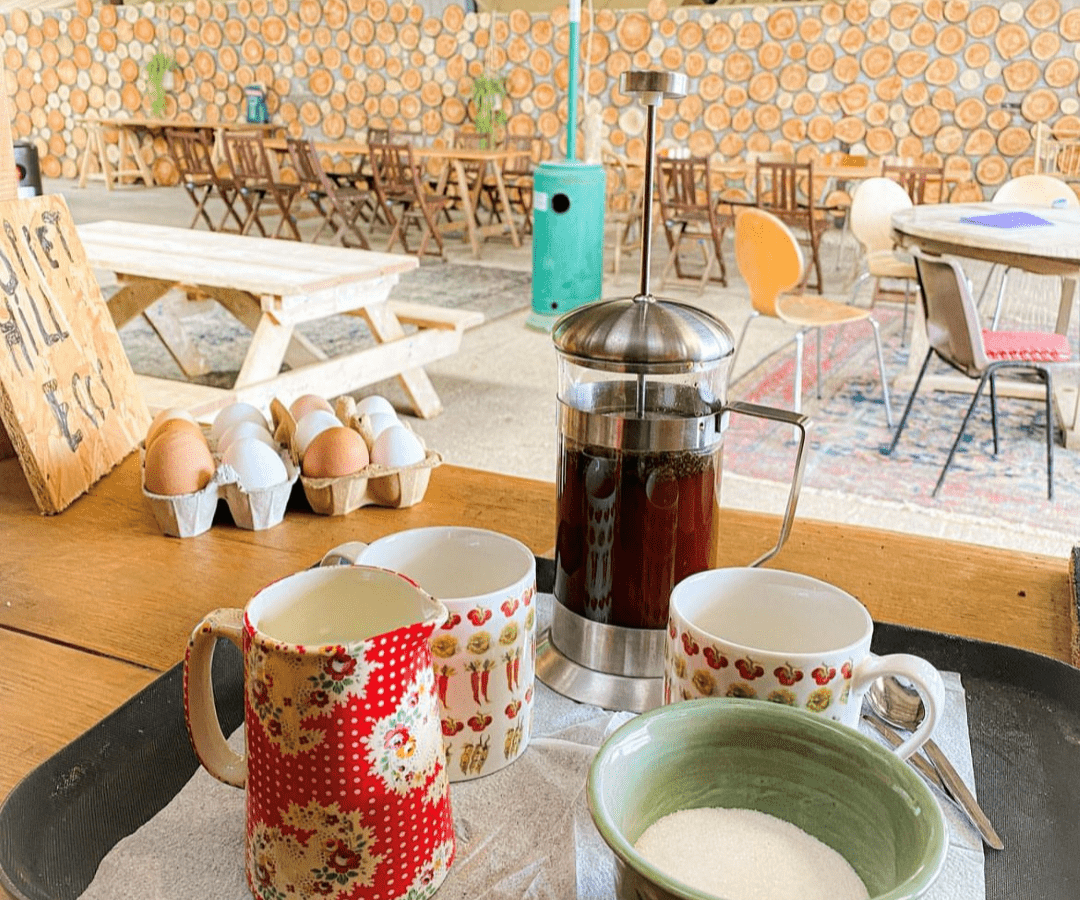 A table with a French press of coffee, two ceramic mugs, a bowl of sugar, and a patterned jug on a tray in the foreground. In the background, Coppet Hill's spacious indoor seating area features wooden tables, chairs, a stack of firewood, and various decorative items.