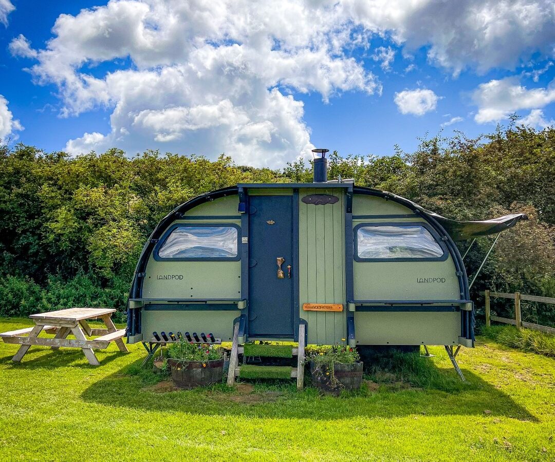 A green, curved wooden cabin stands on grassy land under a partly cloudy sky on Coppet Hill. It has a central door, windows on each side, and a small chimney. A picnic table with benches sits to the left, and lush green trees surround the background.