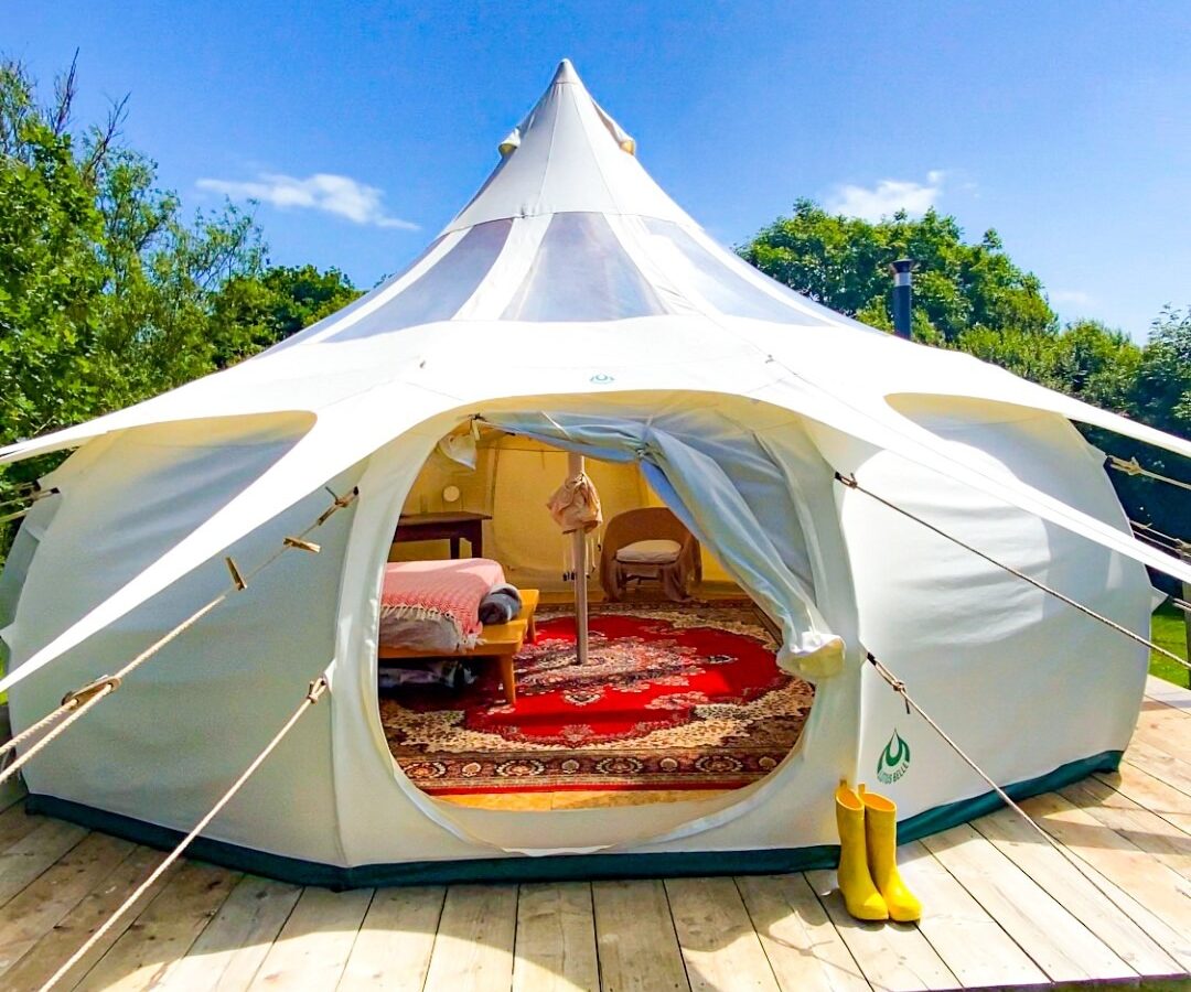 A white canvas tent on a wooden platform sits under a bright blue sky at Coppet Hill. The tent's entrance is open, revealing a cozy interior with a red rug, a small table, and a chair. Yellow rubber boots are placed outside on the grass beside the platform. Lush greenery surrounds the tent.